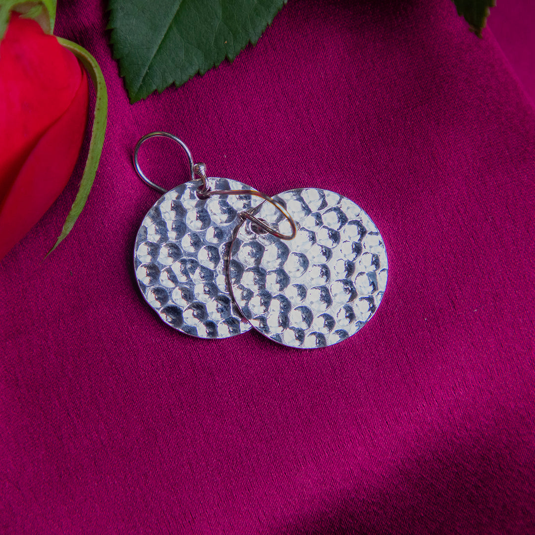A pair of round silver earrings with a tribal hammered pattern, displayed on a pink background with a red rose and green leaves.