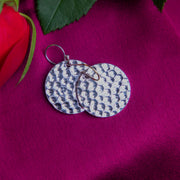 A pair of round silver earrings with a tribal hammered pattern, displayed on a pink background with a red rose and green leaves.