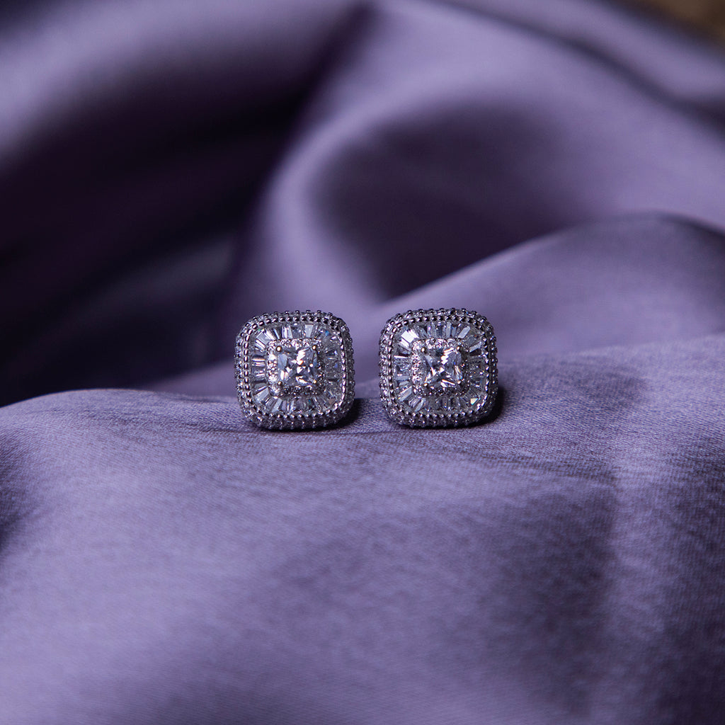 A pair of sterling silver stud earrings with a brilliant-cut white stone, set within a sterling silver bezel, displayed on a purple fabric surface.