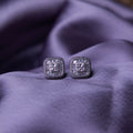 A pair of sterling silver stud earrings with a brilliant-cut white stone, set within a sterling silver bezel, displayed on a purple fabric surface.