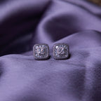 A pair of sterling silver stud earrings with a brilliant-cut white stone, set within a sterling silver bezel, displayed on a purple fabric surface.