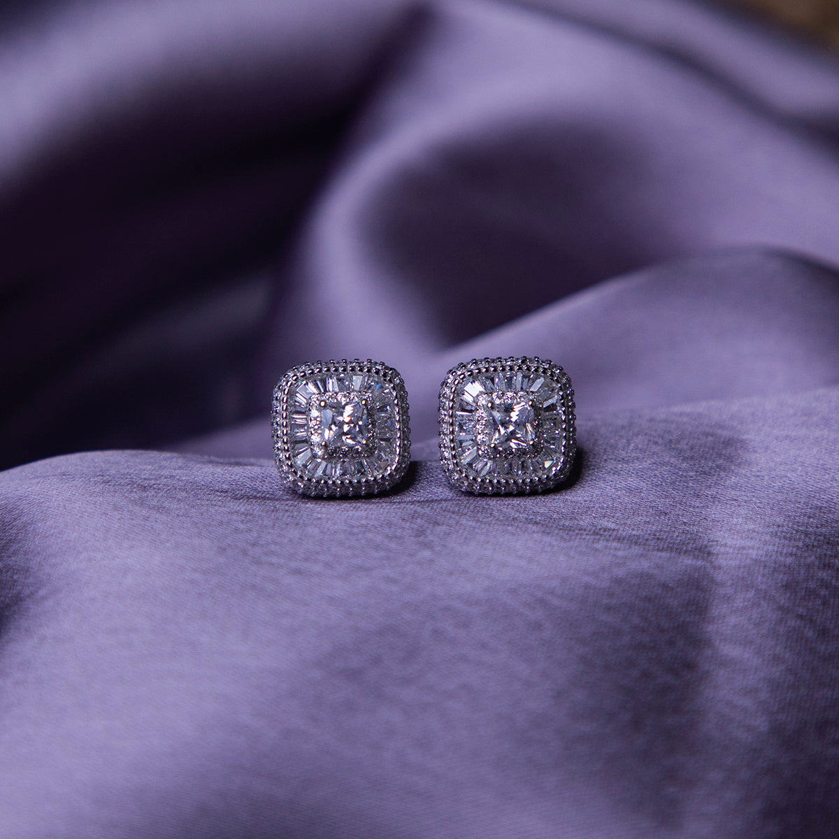 A pair of sterling silver stud earrings with a brilliant-cut white stone, set within a sterling silver bezel, displayed on a purple fabric surface.