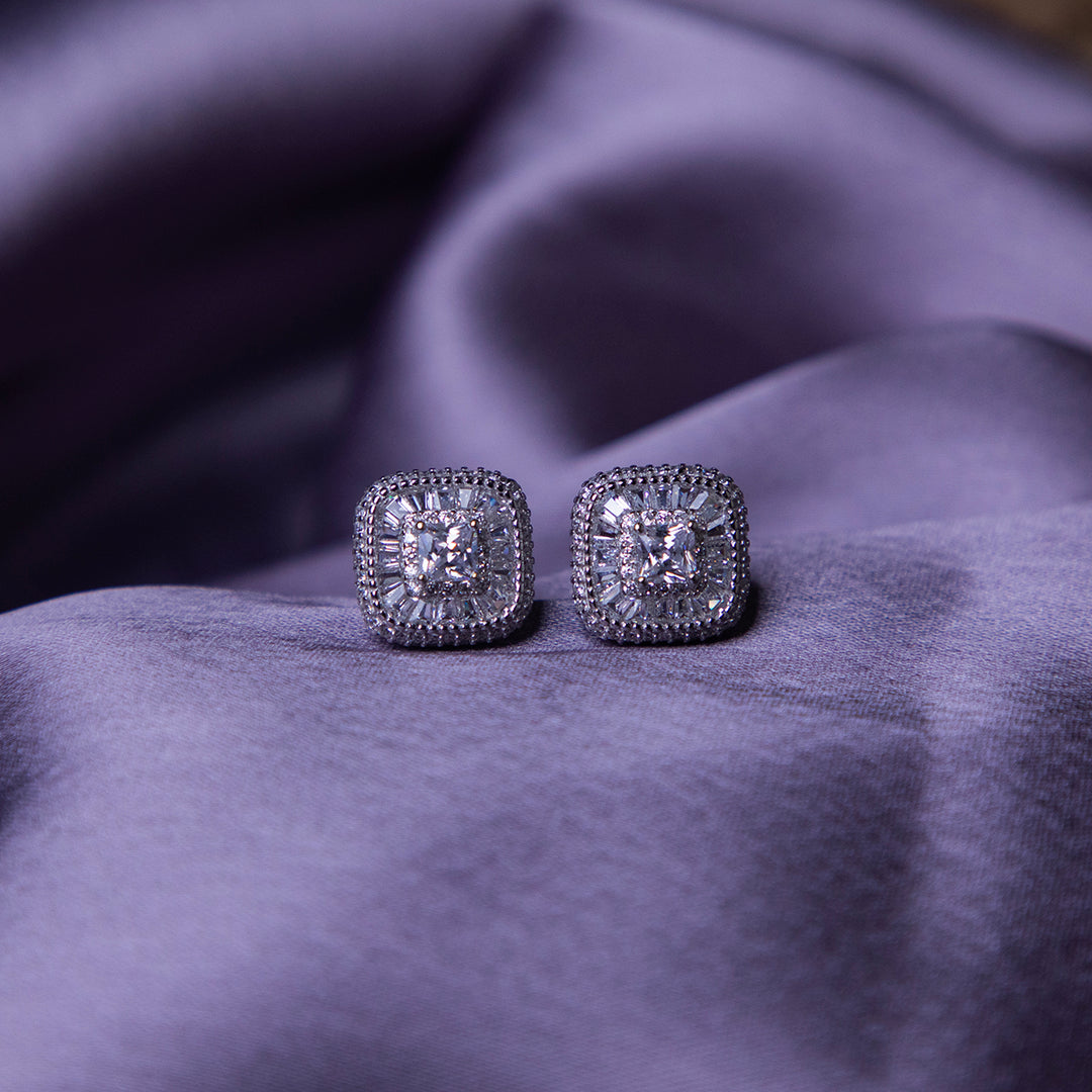 A pair of sterling silver stud earrings with a brilliant-cut white stone, set within a sterling silver bezel, displayed on a purple fabric surface.