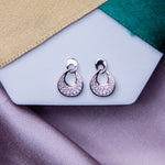 A pair of silver embellished earrings displayed on a white backkground with green and beige ribbon.