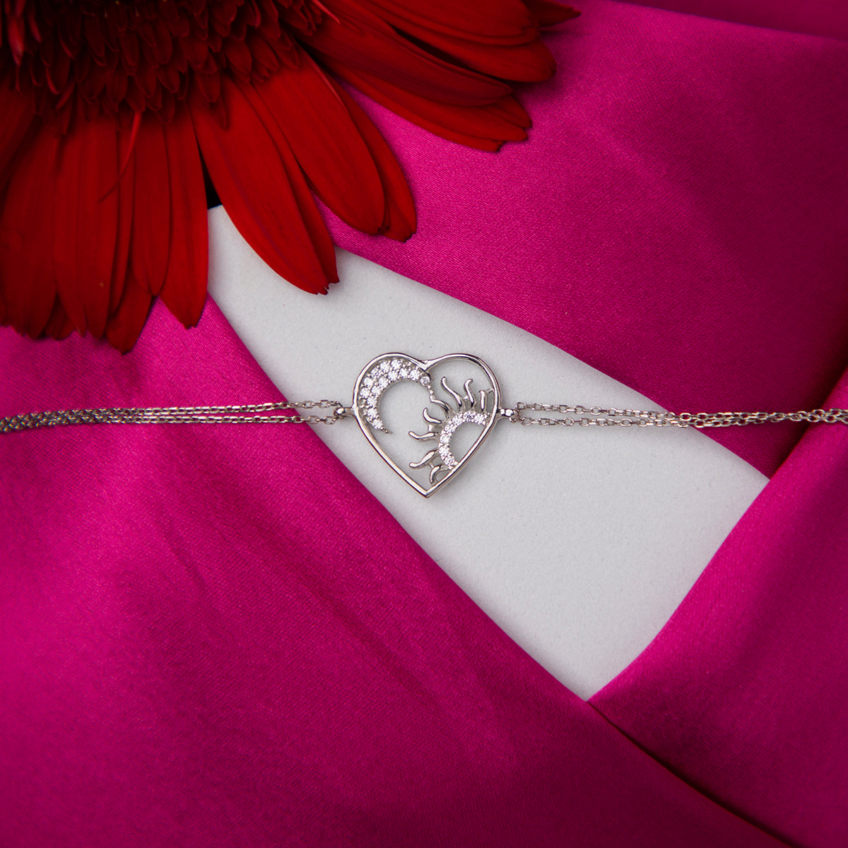 A silver bracelet with a heart-shaped sun charm and a deer head charm, displayed on a wrist against a pink floral background.