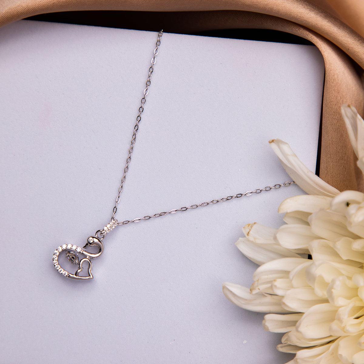A silver pendant chain necklace with a swan-shaped pendant with dancing stone, displayed on a white surface next to a flower.