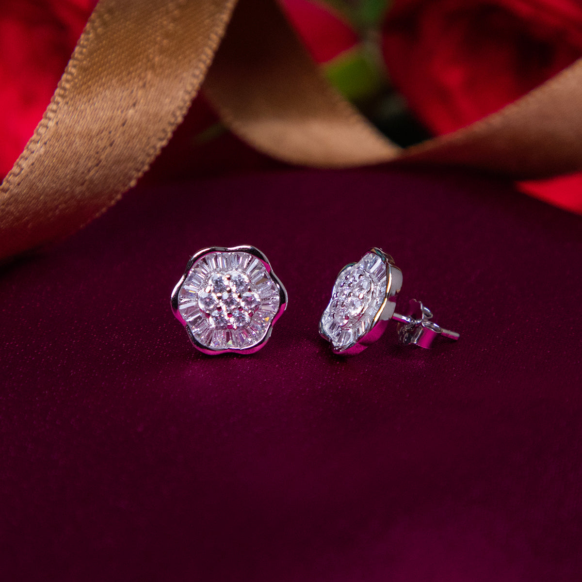 A pair of sterling silver stud earrings with a rhodium-based anti-tarnish coating, displayed on a magenta pink surface with a golden ribbon in the background.