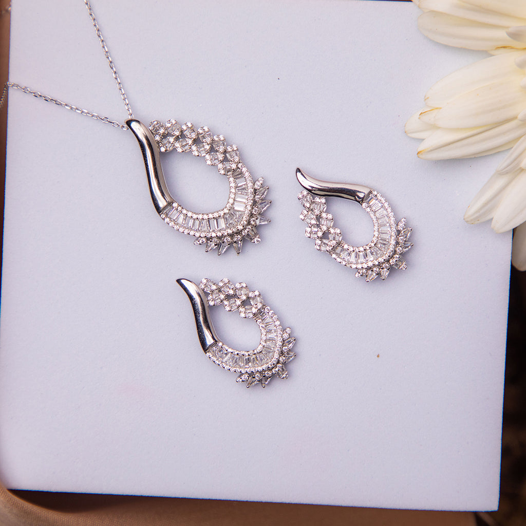 A sterling silver pendant set with white stones accompanied by a pair of earrings, displayed on a white surface next to a flower.
