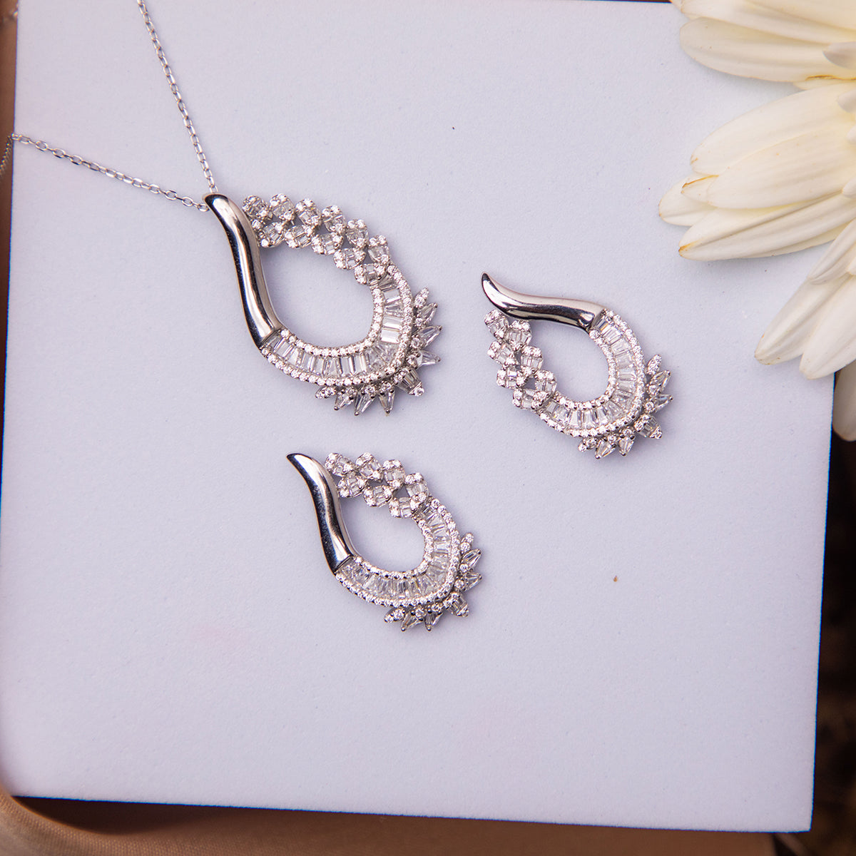 A sterling silver pendant set with white stones accompanied by a pair of earrings, displayed on a white surface next to a flower.