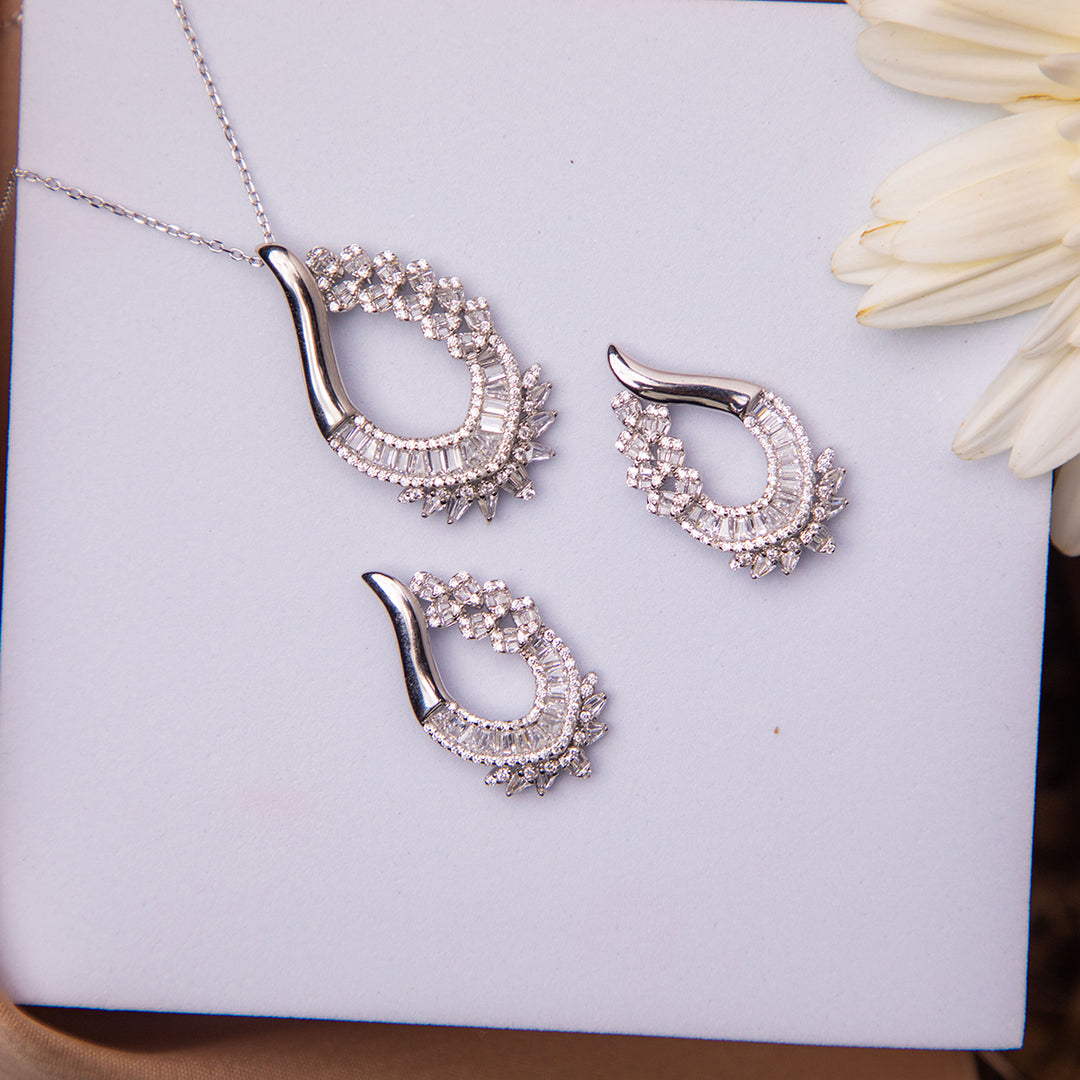 A sterling silver pendant set with white stones accompanied by a pair of earrings, displayed on a white surface next to a flower.