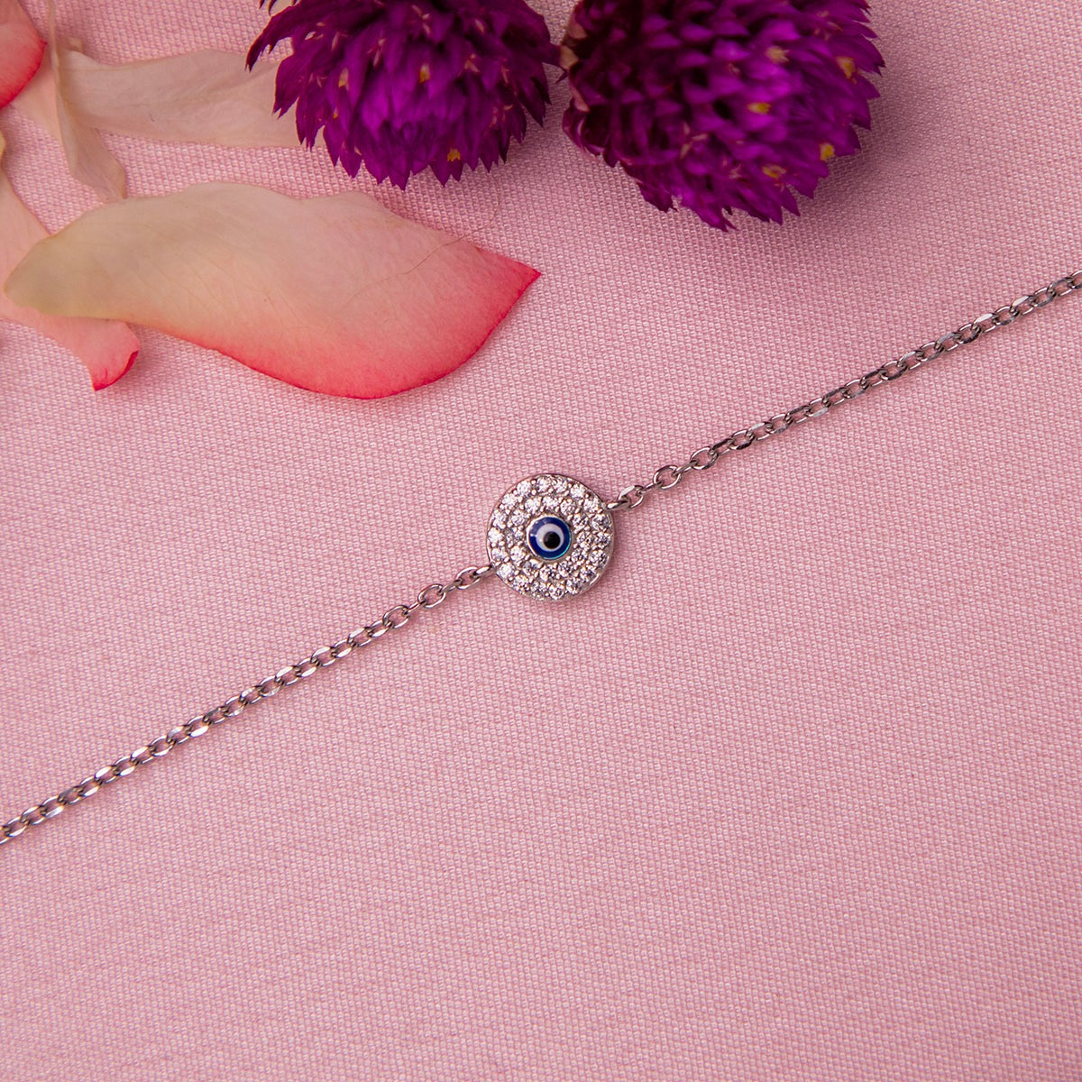 A silver-colored bracelet with a blue evil eye charm, displayed on a pink background with a purple flower and pink petal in the corner.