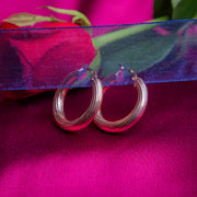 A pair of rose gold-colored hoop earrings placed on a purple velvet surface with a red rose in the background.