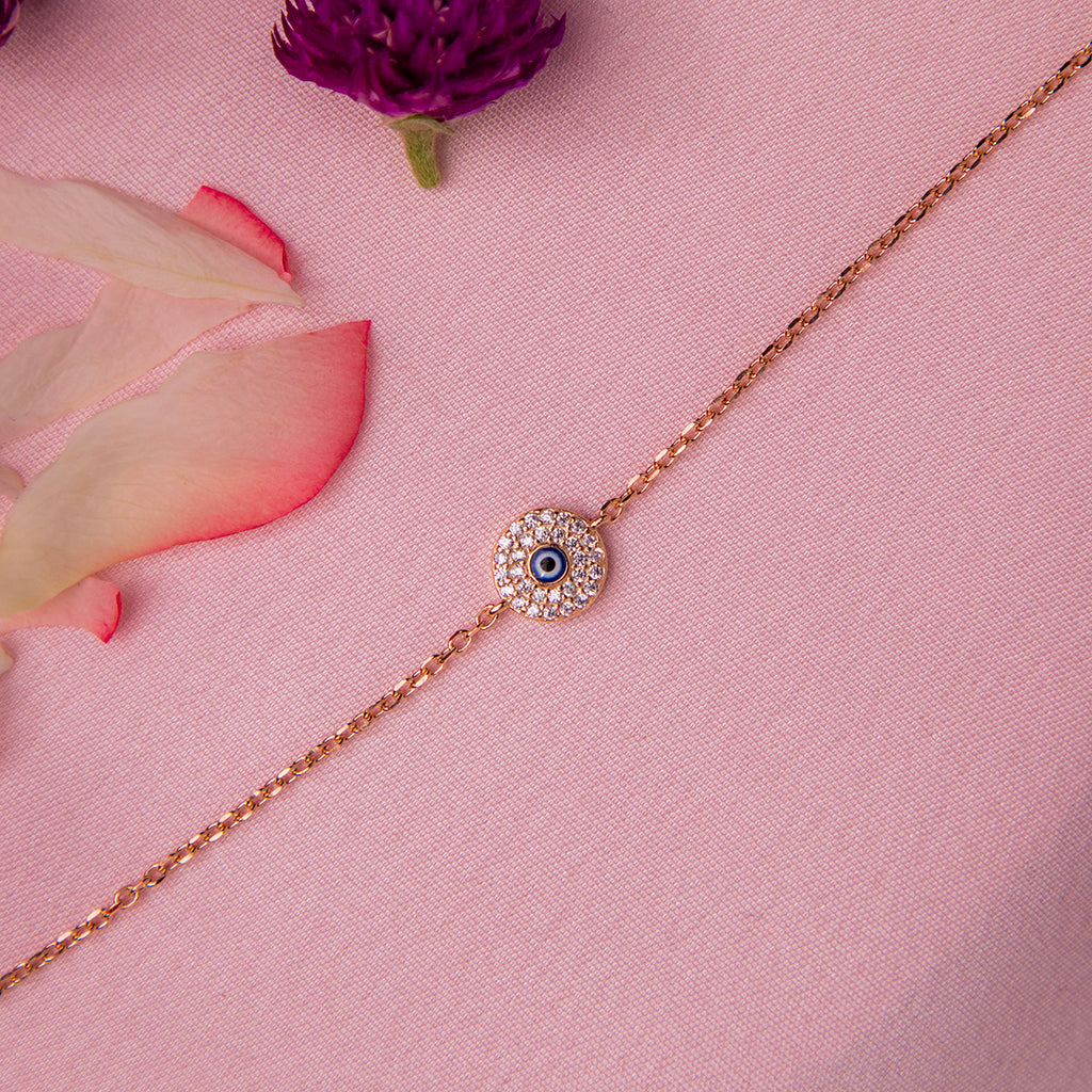 A rose gold-colored evil eye bracelet with a crystal-encrusted motif on a pink background, surrounded by white and pink flower petals.