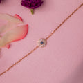 A rose gold-colored evil eye bracelet with a crystal-encrusted motif on a pink background, surrounded by white and pink flower petals.