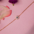 A rose gold-colored evil eye bracelet with a crystal-encrusted motif on a pink background, surrounded by white and pink flower petals.