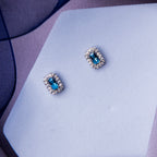 A pair of turquoise power embellished stud earrings placed on a white surface with a blue textured background.