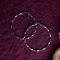 A pair of silver hoop earrings with a twisted contemporary design, placed on a textured purple surface.