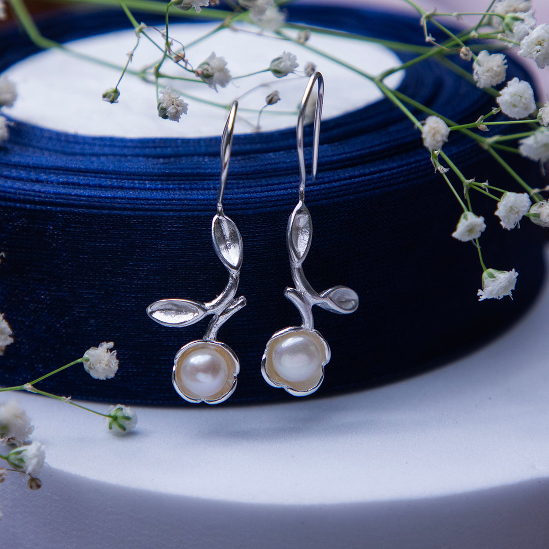 A pair of sterling silver pearl earrings with rose-inspired accents, displayed on a blue spool of thread with white flowers around it.