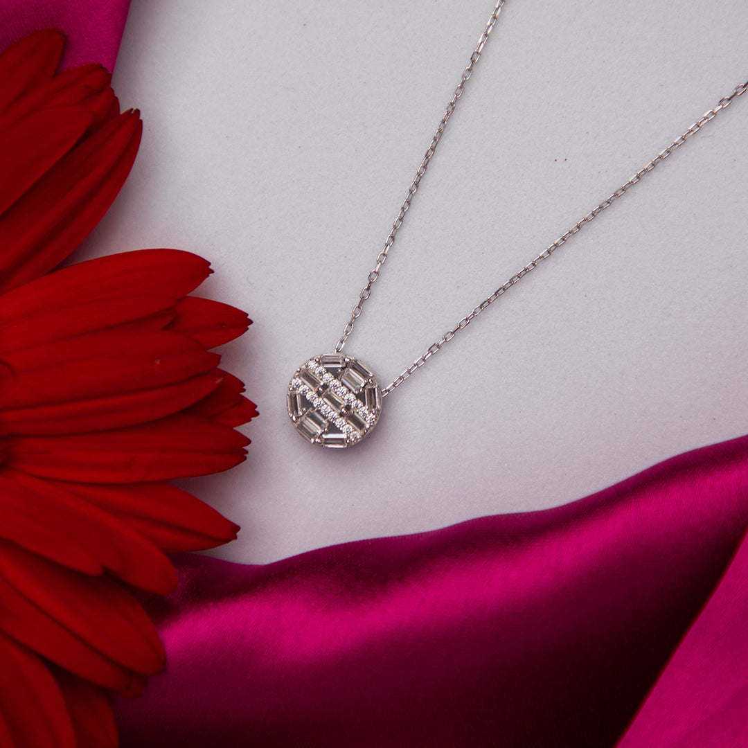 A sterling silver pendant chain with a round white stone pendant, displayed on a pink fabric surface with a red flower nearby.