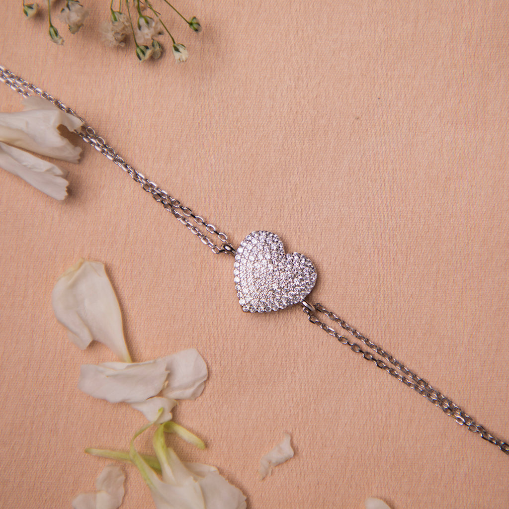 A silver bracelet featuring a heart-shaped charms with white stone studs, displayed on a neutral background with flower petals scattered around.