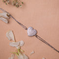 A silver bracelet featuring a heart-shaped charms with white stone studs, displayed on a neutral background with flower petals scattered around.
