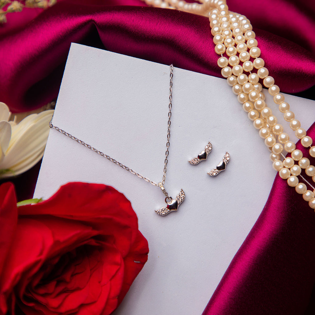 A sterling silver pendant set featuring heart-shaped pendants with wings, accompanied by a pair of earrings, displayed on a white card with a red rose and pearls in the background.