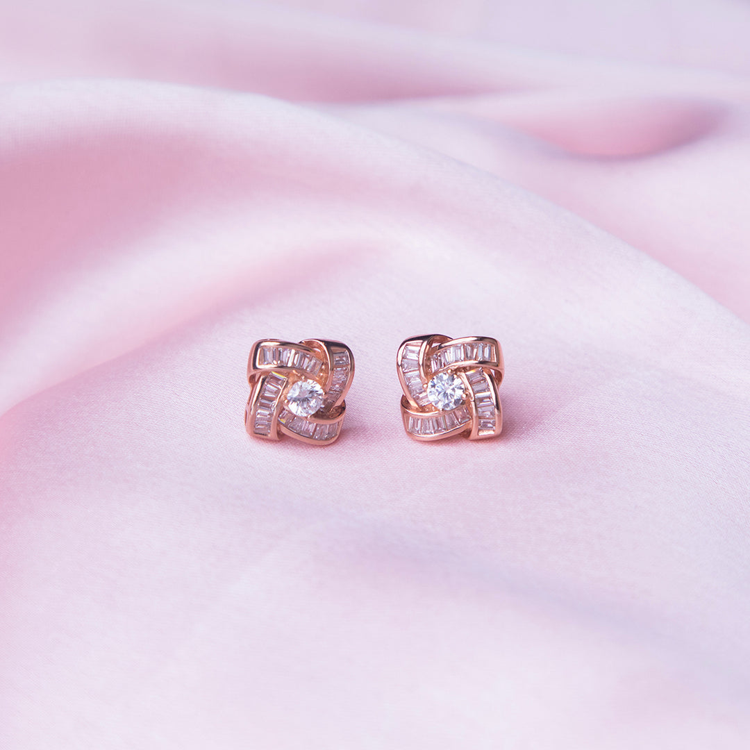 A pair of rose gold-colored silver stud earrings with white stones set in a geometric pattern, displayed on a pink background.